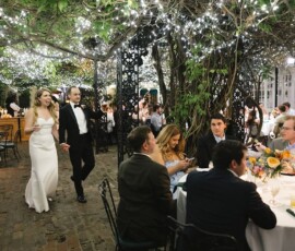 Bride and groom walk hand in hand at an outdoor Gulf South wedding under string lights, with guests among lush greenery.