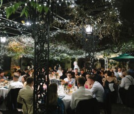 Guests in formal attire mingle at round tables under string lights, enjoying a festive New Orleans wedding outdoors.