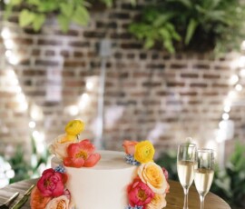 White frosted cake with colorful flowers on a wooden table beside champagne glasses, brick wall and string lights in background.
