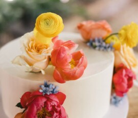 White frosted cake with pink peonies, yellow ranunculus, peach roses, and blue blossoms on wood—ideal for New Orleans wedding.