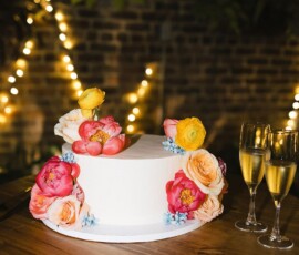 Heart-shaped white cake with colorful flowers beside two champagne glasses, string lights, and greenery in the background.