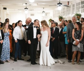 Bride and groom celebrate their indoor wedding reception, surrounded by formally dressed guests in a crowded room.