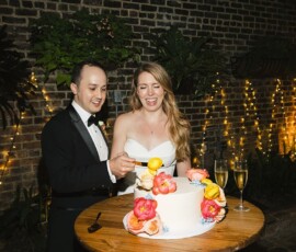 Smiling couple in formalwear cuts a floral wedding cake at their Louisiana wedding, with champagne nearby and string lights behind.