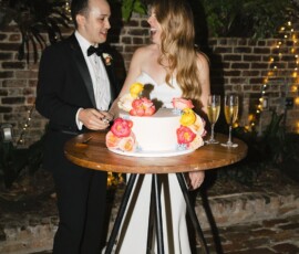 Formal couple by a wedding cake and champagne flutes, with string lights on a brick wall at a classic New Orleans wedding.