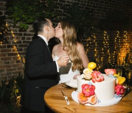 A couple in wedding attire kisses by a decorated cake, with string lights and greenery at their New Orleans wedding.