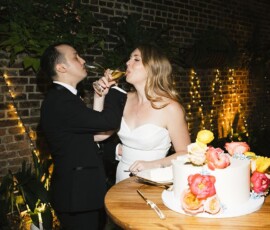 Bride and groom toast with champagne beside wedding cake at elegant New Orleans wedding with string lights and greenery.