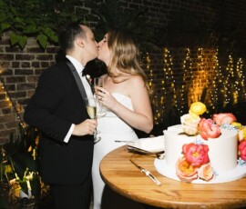 Newlyweds kiss holding champagne by a floral wedding cake, with string lights and plants on a brick wall backdrop.