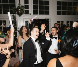 Louisiana wedding party: two men in tuxedos dance, smiling, amid costumed guests in a bright room with large windows.
