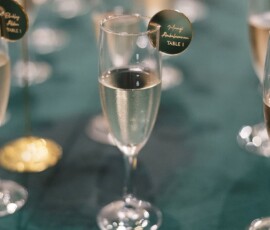 Champagne glass on green tablecloth with “Happy Anniversary TABLE 1” tag; blurred glasses in background.