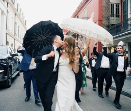 Bride and groom kiss as they lead a joyful New Orleans wedding parade; guests follow with umbrellas, celebrating.