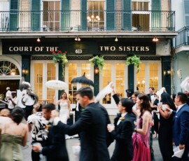 Bride with white parasol leads festive wedding parade outside Court of the Two Sisters; guests and musicians celebrate.