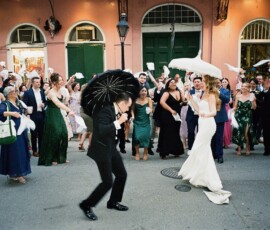New Orleans wedding parade with a dancing bride and groom holding umbrellas, joined by cheering guests waving handkerchiefs.
