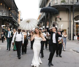 Bride in white dress leads musicians and guests down city street, smiling with a parasol and festive wedding energy.