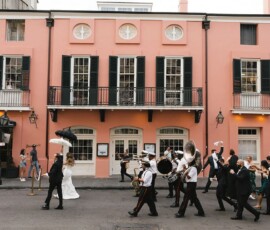 Bride and groom lead a lively brass band parade past a pink building, followed by cheering guests at a New Orleans-inspired wedding.