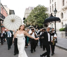 Bride and groom lead a joyful Louisiana wedding parade with parasols, musicians, guests, and historic city buildings.