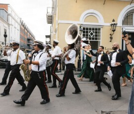 A marching band leads a wedding procession with bride and groom on a city street, as a woman takes photos for inspiration.