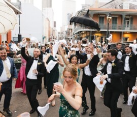 Wedding parade in New Orleans with guests dancing in formal wear, waving napkins, surrounded by musicians and umbrellas.