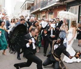 A lively New Orleans wedding parade with a groom’s black umbrella, dancer in sash, and guests celebrating with parasols.