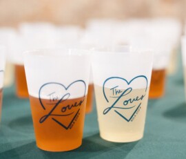 Two drinks in plastic cups with “The Loves EST. 2022” in blue hearts on a green table—wedding inspiration.
