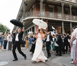 Bride and groom lead a festive Louisiana street parade with umbrellas, joined by cheering guests waving handkerchiefs.