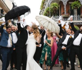 Bride and groom kiss in the street as a joyful Gulf South wedding party celebrates with parasols, handkerchiefs, and lush greenery.