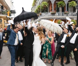 Bride and groom kiss in the street as guests with umbrellas cheer, with Louisiana balconies and plants in the background.