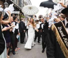 Bride and groom parade joyfully down a lively New Orleans street, with guests waving napkins and musicians leading the way.