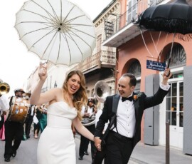 Bride and groom parade down a city street with umbrellas and a brass band, celebrating their Gulf South wedding.