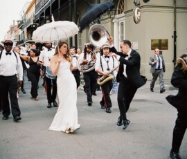 Bride and groom lead a lively brass band parade with guests, umbrellas, and musicians at a festive Louisiana wedding.
