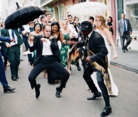Formally dressed people dance joyfully with umbrellas in a festive New Orleans street, city buildings in the background.