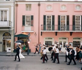 A joyful Louisiana wedding parade with a bride, musicians, pastel buildings, onlookers, and umbrellas lining the street.
