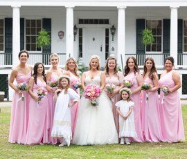 Bride, nine bridesmaids in pink, and two flower girls in white pose with flowers by a grand white-columned Louisiana house.