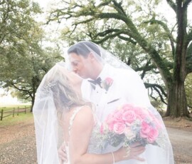 Bride and groom kiss under a veil outdoors, pink rose bouquet in hand, trees and a fence behind at their New Orleans wedding.