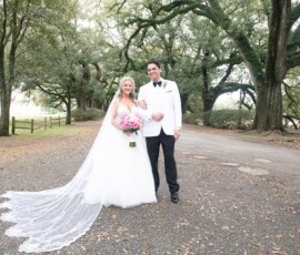 Bride in white gown and groom in white tuxedo smile on a tree-lined path, bride holding pink bouquet—Gulf South wedding inspo.