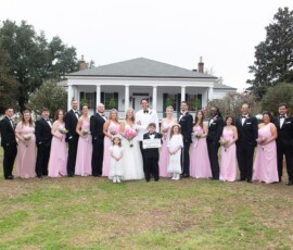 Bride, groom, bridesmaids in pink, groomsmen in black, and children pose smiling outside a white house at a Gulf South wedding.