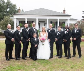Bride in white gown with young boy and nine groomsmen pose on lawn before a columned white house, New Orleans wedding style.