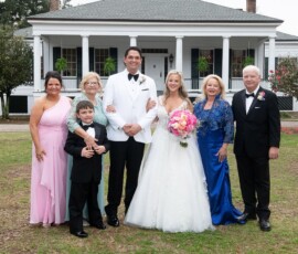 Bride, groom, four adults, and a child pose formally outside a columned white house for a Louisiana wedding photo.