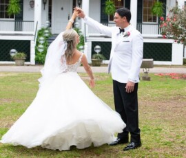 Bride and groom smile, holding hands as they dance outside a white house at their Louisiana wedding.