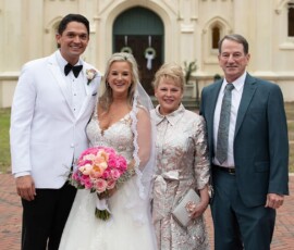 Bride and groom with an older couple, all smiling in formal wear outside a cream church; bride holds pink and white bouquet.