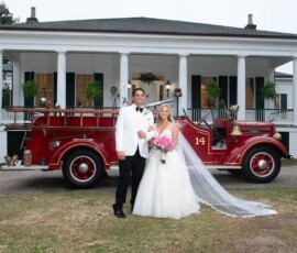 Bride and groom by a vintage red fire truck outside a white-columned house, her pink bouquet and veil adding New Orleans wedding charm.