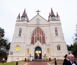 White Gothic church with red spires at dusk; guests in formal attire gather for a Louisiana wedding, palms and trees flank the entrance.
