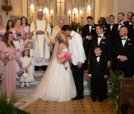Bride and groom kiss at altar, surrounded by bridal party, priests, and ring bearer in beautiful Gulf South church wedding.