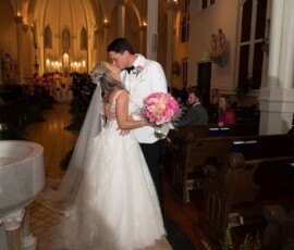 Bride and groom kiss at the altar, pink bouquet in hand, with guests seated in warmly lit church with arches and candles.