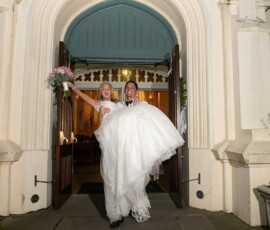 Groom in tuxedo carries smiling bride in white dress and bouquet outside arched doors, classic New Orleans wedding.