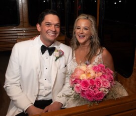 Smiling bride in white dress with pink bouquet and groom in white tuxedo sit on a trolley’s wooden bench.