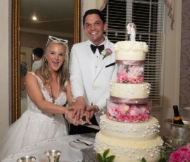 Smiling bride and groom cut a white, three-tiered cake with pink flowers at their Louisiana wedding. Champagne nearby.