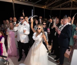 Bride in white gown and heart-shaped glasses celebrates with guests waving sparklers at a lively night wedding reception.