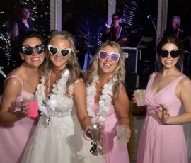 Four women in pink dresses and sunglasses pose at a Louisiana wedding; the bride holds a trophy, live band behind them.