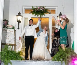Bride in white dress and jean jacket, groom in tux celebrate on porch with silly string; wedding sign displayed.