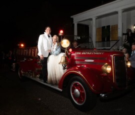 Bride and groom smile on a vintage red fire truck at night, with guests and a white-columned building in the background.